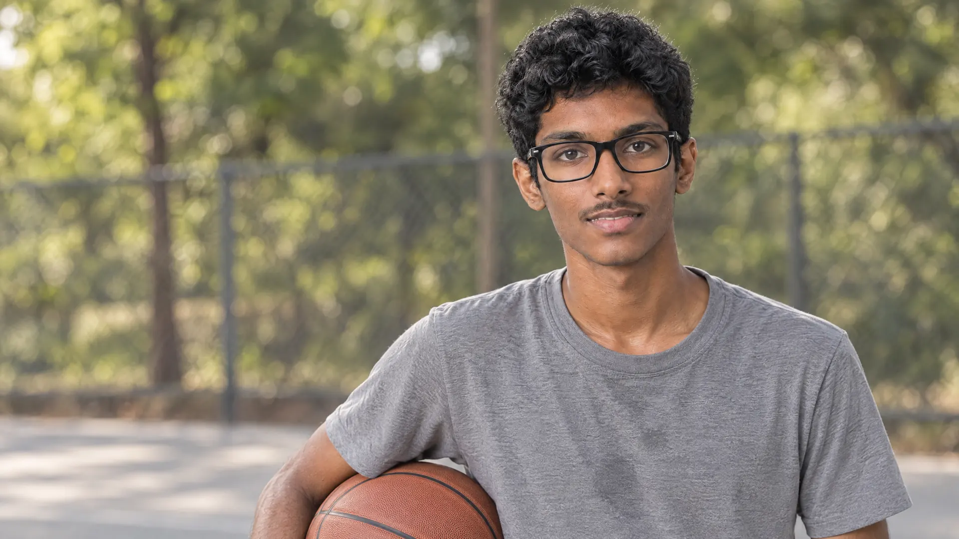 A young man of about nineteen with short curls and bold black glasses holds a basketball on an outdoor court in daylight, his lenses clean and unmarked — the hard coat.