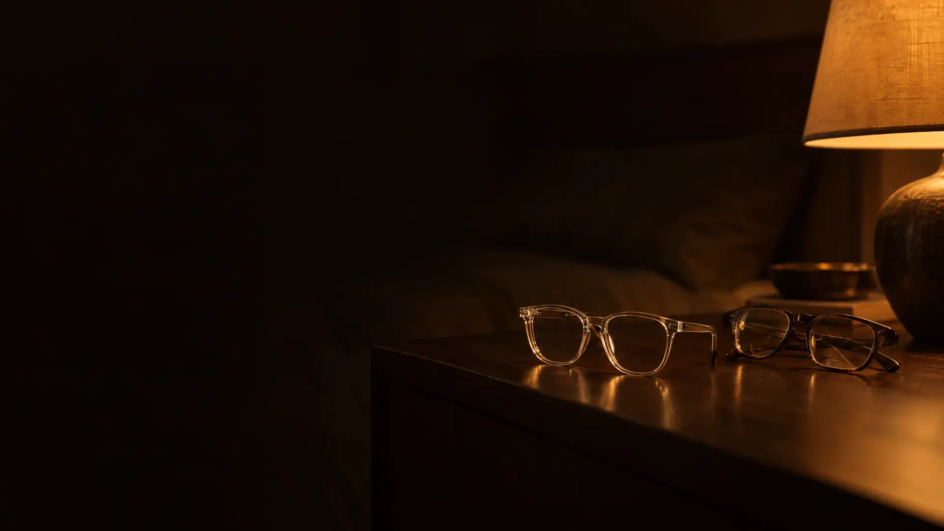 A new pair and an old pair of glasses side by side on a bedside table at night.