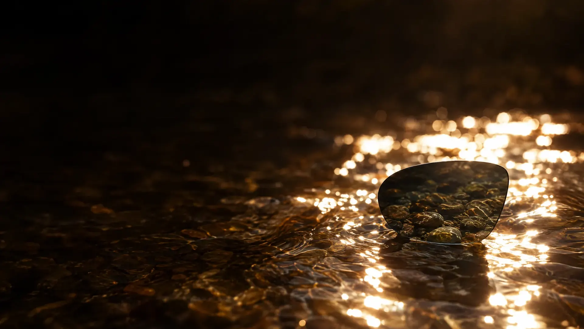 A dark lens above sunlit water, the glare gone through it and the pebbles below clear.