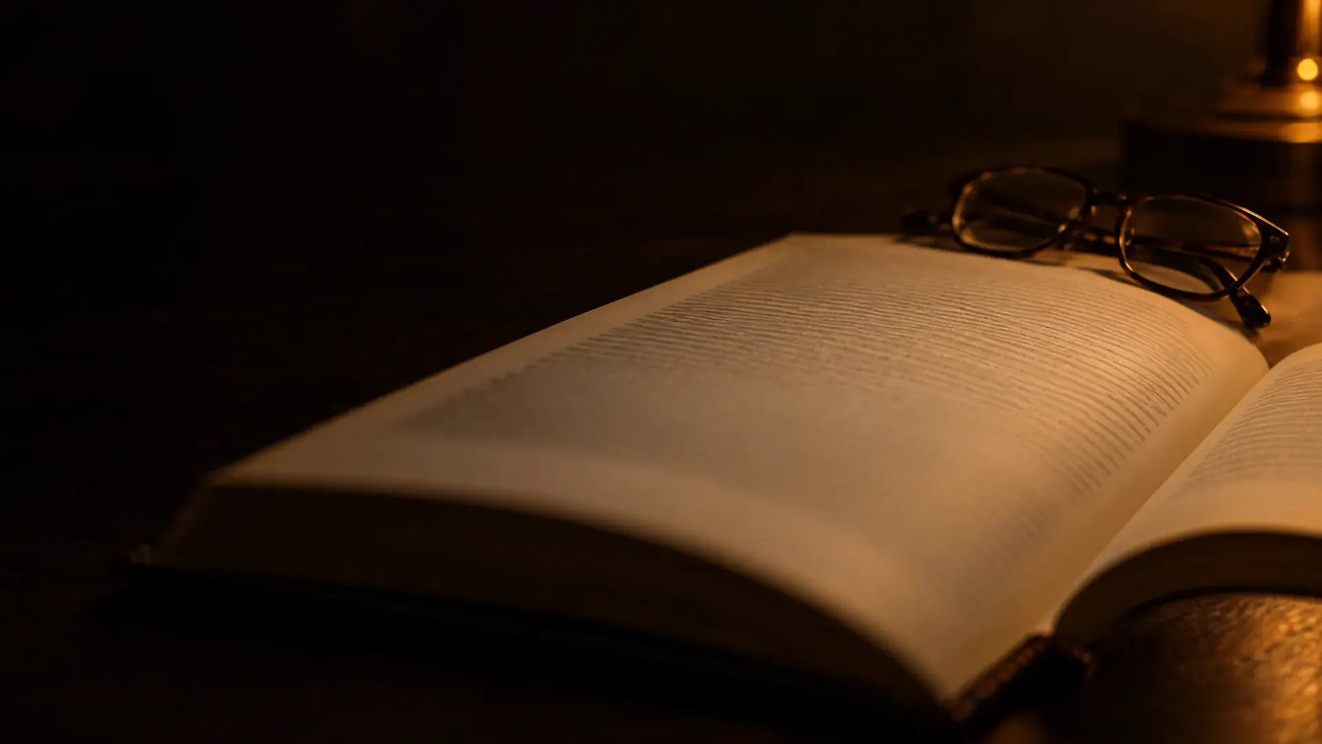 An open book on a dark desk in lamplight, its nearest page soft and out of focus, reading glasses at the far edge.