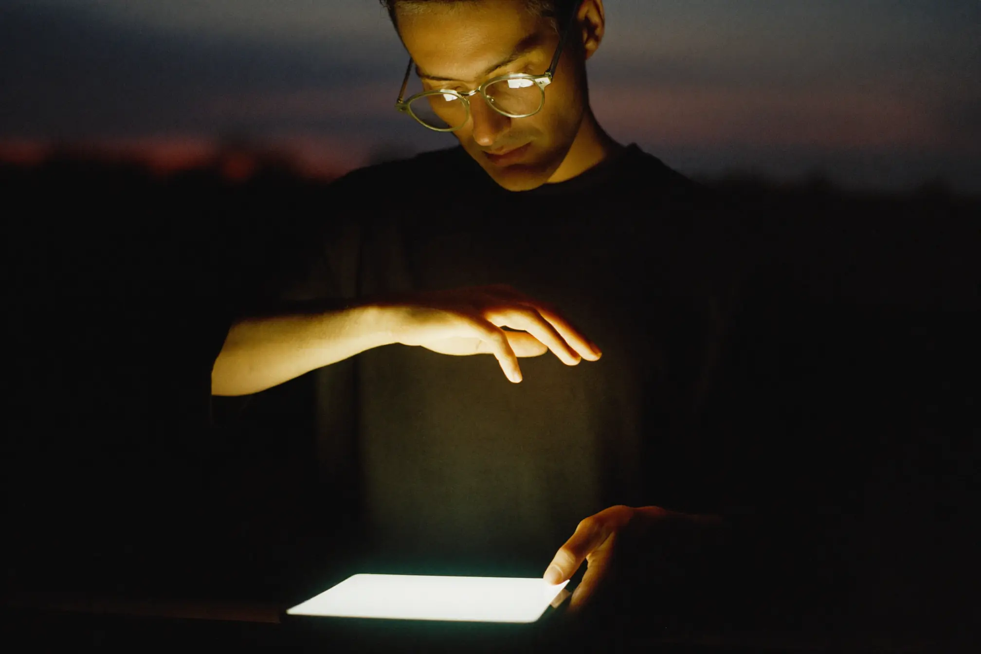 A founder standing at a dark window at dusk with a soft warm-white tablet glow.