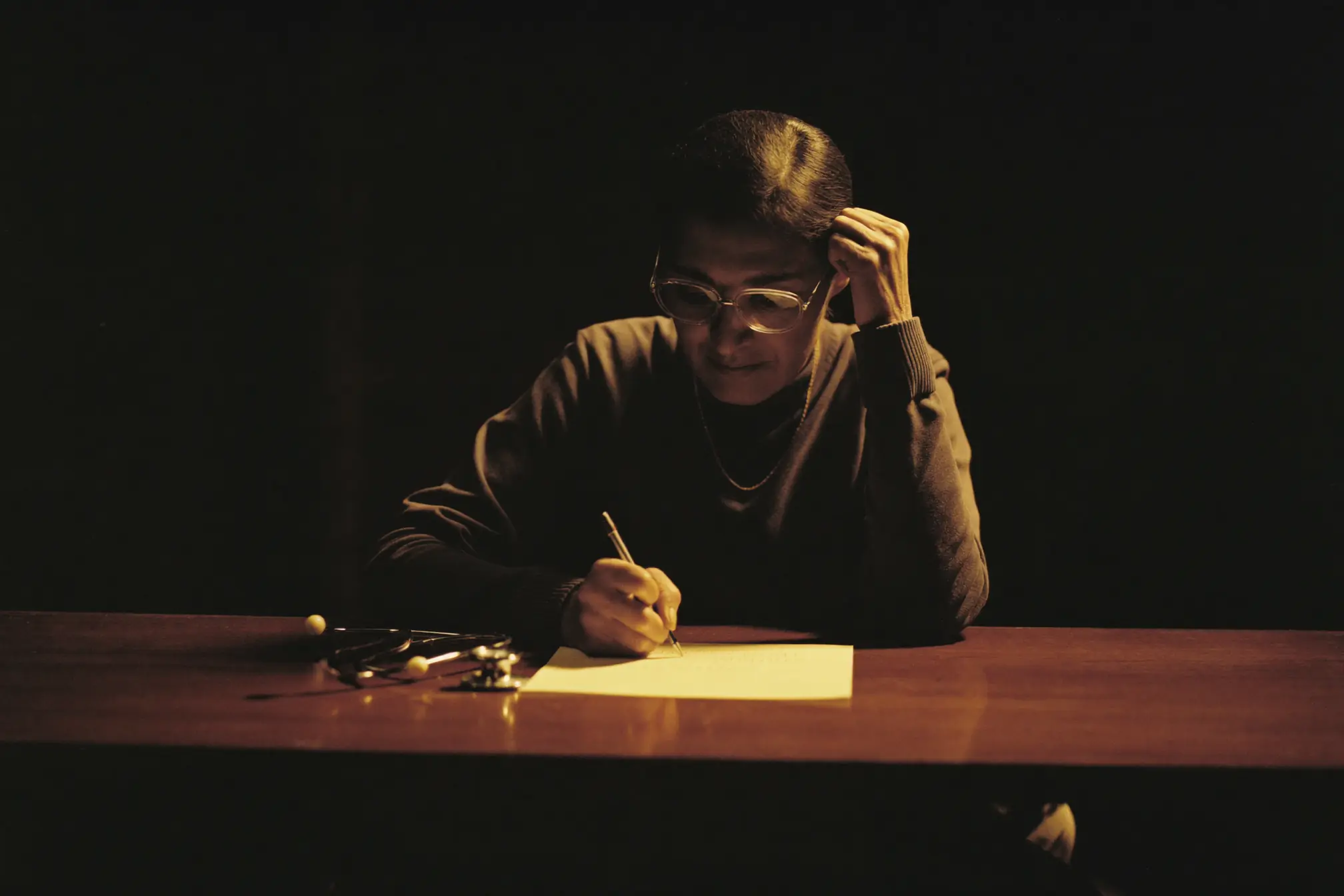 A primary care physician writing on a blank pad at a dark wooden desk.