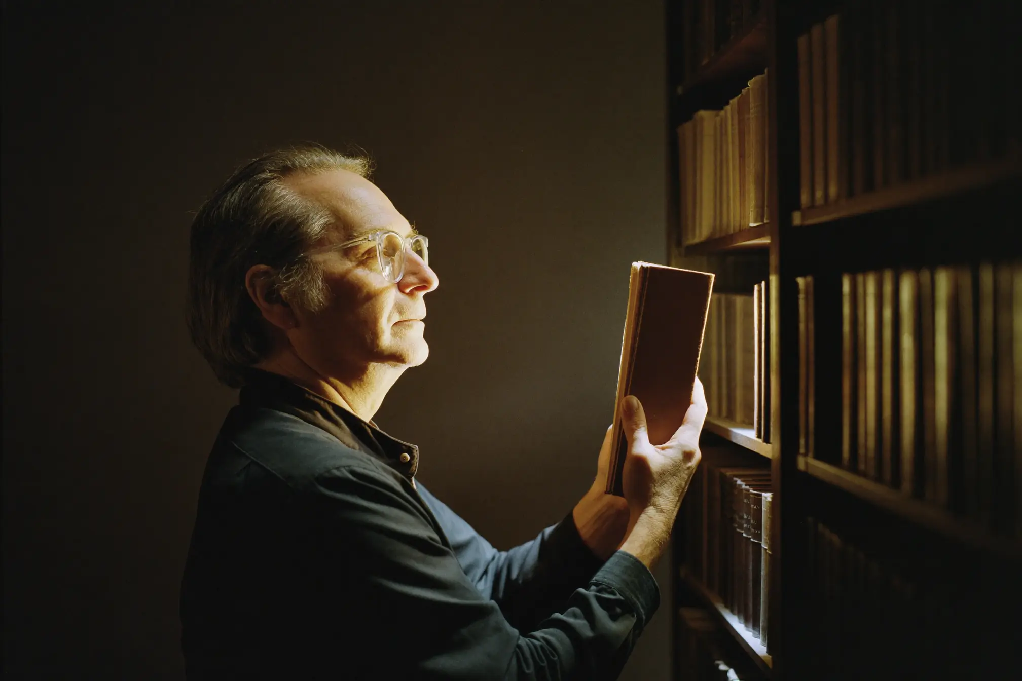 A librarian holding an old bound volume close in a dark aisle of shelves.