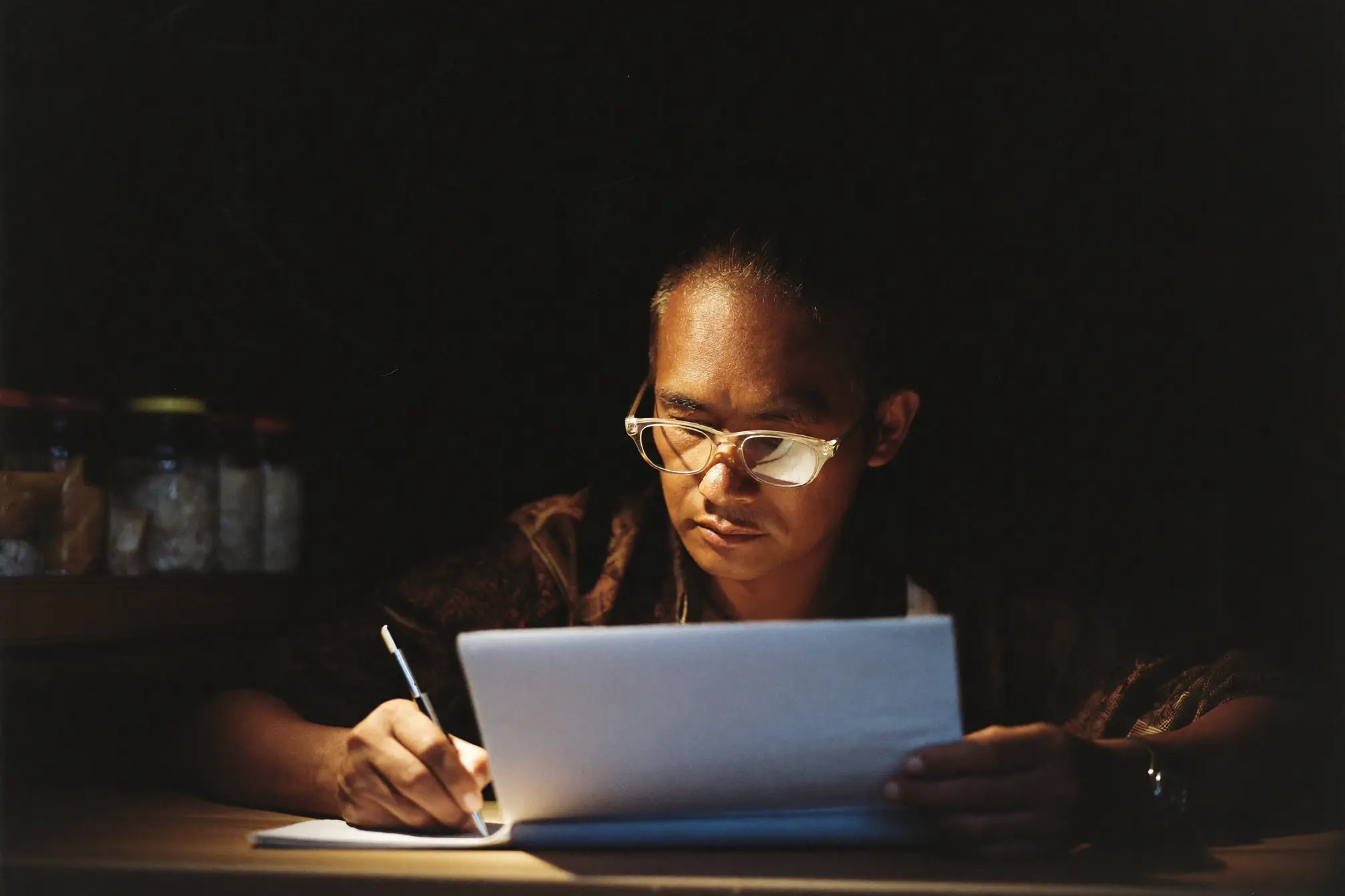 A small shop owner noting stock in a blank register at a wooden counter.