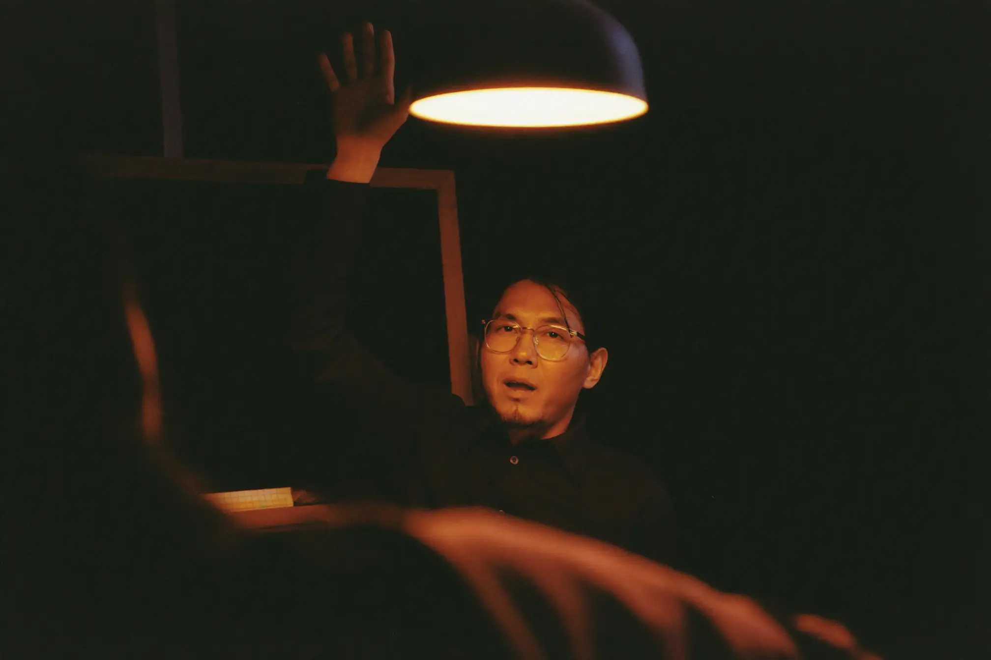 A private tutor explaining with a raised hand beside a small dark chalkboard with nothing written on it.