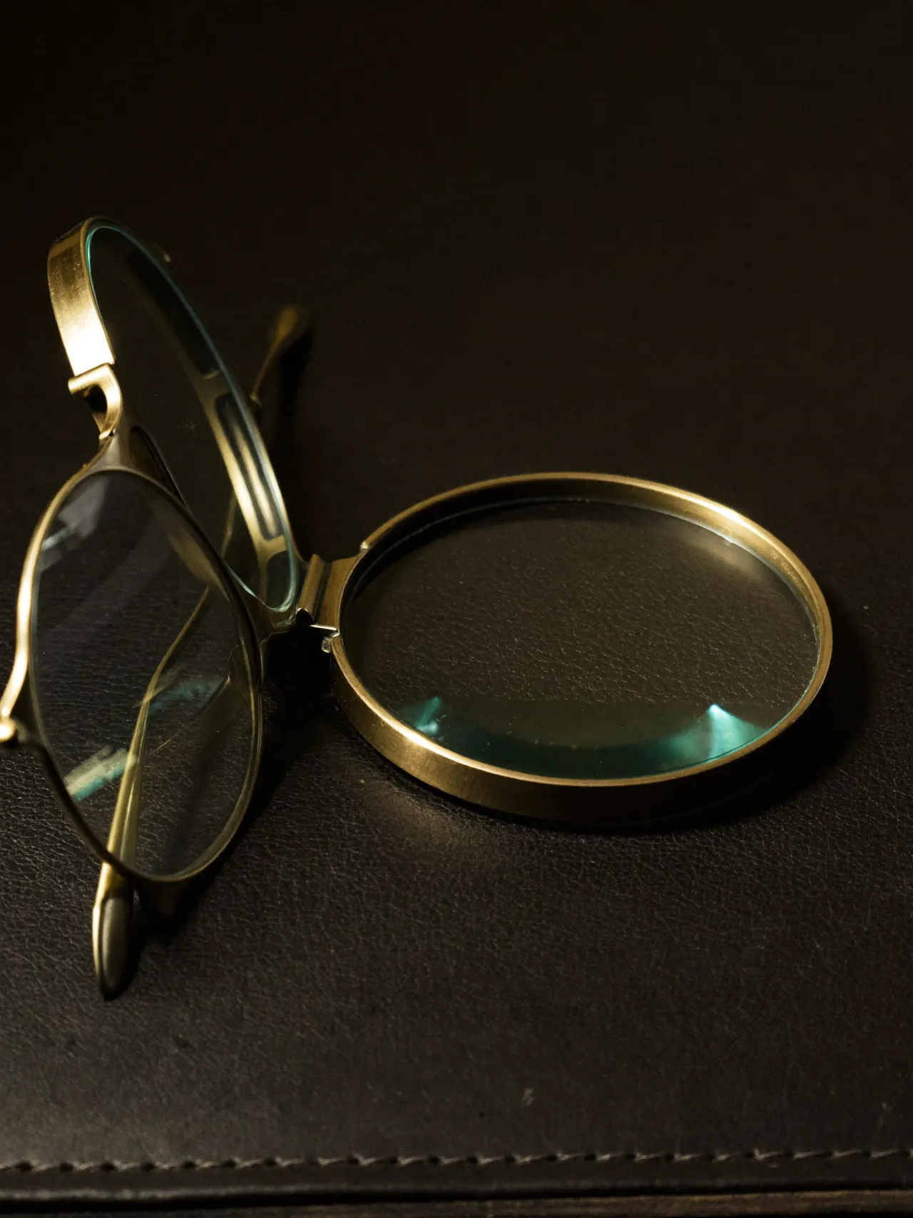 A brass loupe lying open beside a pair of thin metal frames on a dark leather counter, lit from the left.