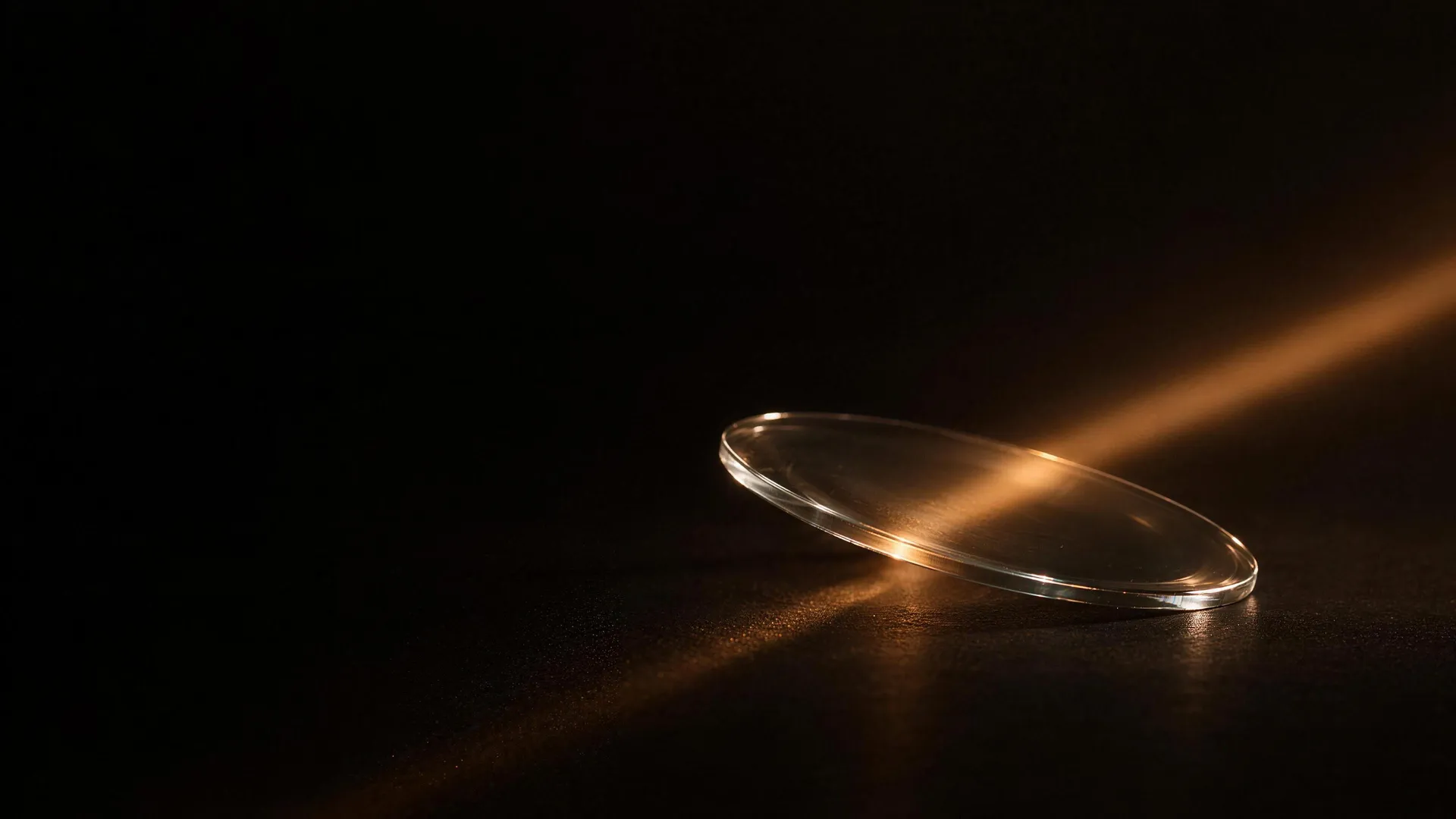 A clear lens standing on a dark surface, lit by a single narrow beam of warm light that crosses it and catches its polished rim.