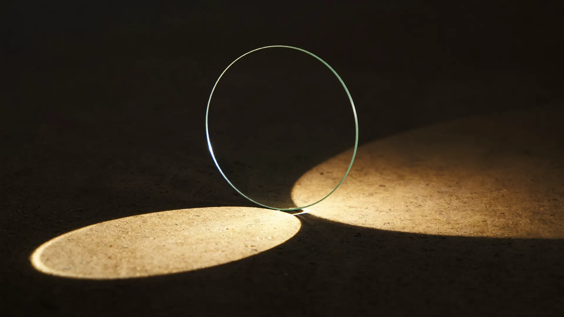 A disc of clear optical glass standing on edge on dark stone, a beam of warm light passing through it and casting a bright oval of light beside it.